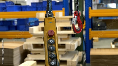 Industrial yellow crane remote control pendant hanging next to a heavy duty red lifting hook in a manufacturing factory warehouse with pallets in the background