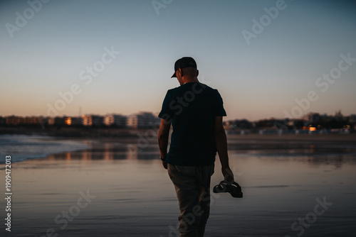 Φωτογραφία A man walks along a quiet, wet shoreline at dusk, holding a pair of sneakers