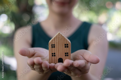 Woman holding a small wooden house in her hands outdoors, symbolizing home care, property protection, real estate investment, family security, sustainable living, and future housing concept.