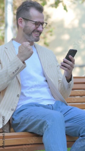 Modern Casual Man Celebrating Success on Smartphone while Sitting Outdoor on a Bench, Vertical Video