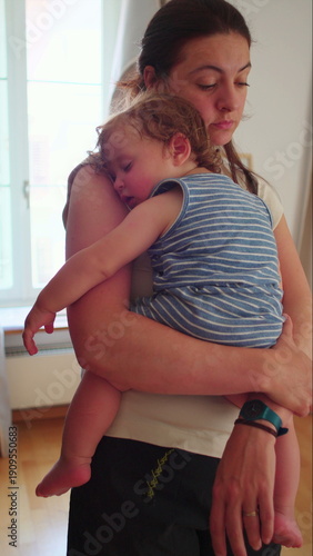 Mother holding sleeping toddler in arms near window, natural light casting soft glow on peaceful embrace during quiet indoor moment