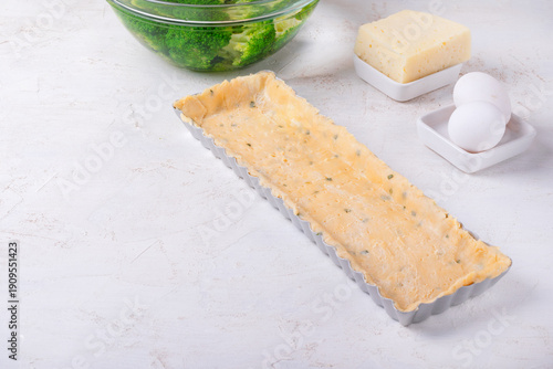 Shortbread pastry dough with herbs in rectangular pan, cheese, chicken eggs, and blanched broccoli on a white table. Step by step cooking tart. Copy space