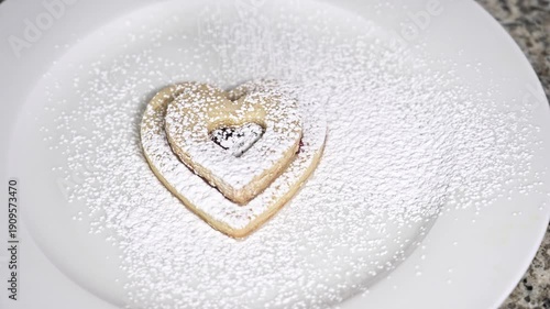Powdered sugar slowly falling over a delicious heart shaped linzer cookie with raspberry jam filling. The sweet dessert sits on a clean white plate