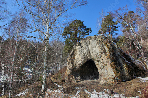 Natural stone cave entrance in a forest landscape with birch trees and melting snow. Early spring tranquil outdoor scene for nature background. Gremyachaya Griva Park, Krasnoyarsk