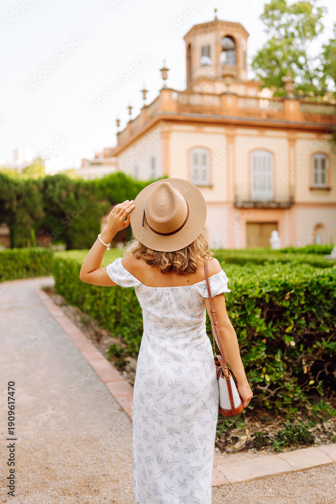 Fototapeta premium A woman stands outdoors in a garden with bushes. She wears a hat and smiles as she adjusts it. A building is visible behind her during late afternoon light.