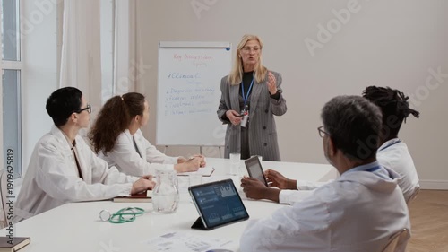 Caucasian blonde mature health professional giving speech on treatment options to multiethnic group of physicians sitting at large table in conference room