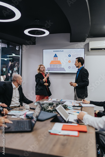 Wallpaper Mural A presenter speaks beside a large screen displaying a business plan info graphic. Colleagues sit around a table with laptops, papers, and coffee in a professional office setting. Torontodigital.ca