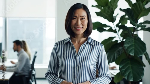 Wallpaper Mural Smiling Asian woman in striped shirt in office setting, plant in background, colleagues blurred Torontodigital.ca