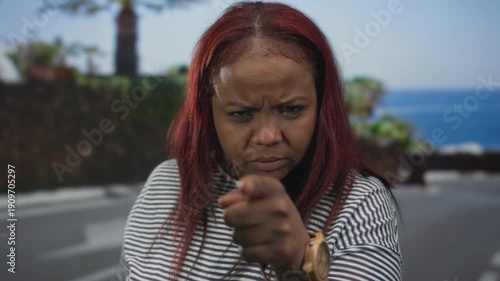 Redheaded african american woman pointing finger at camera on sunny seaside street near ocean promenade; anger.