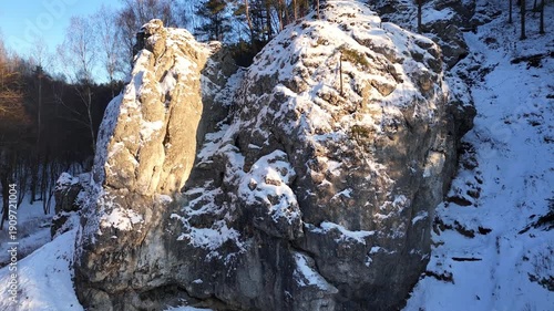 Snow-covered rocky cliff with pine trees and sunlight illuminating the surface, showcasing the gradual transition of light and shadow across the rugged terrain in a winter landscape. Aerial view