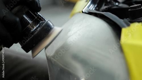 Professional car body shop technician using a pneumatic orbital sander tool for headlight restoration and surface polishing in a modern automotive repair facility