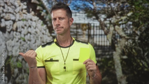 Man points thumb and holds yellow card by stone gate at building entrance; rule enforcement authority sports.