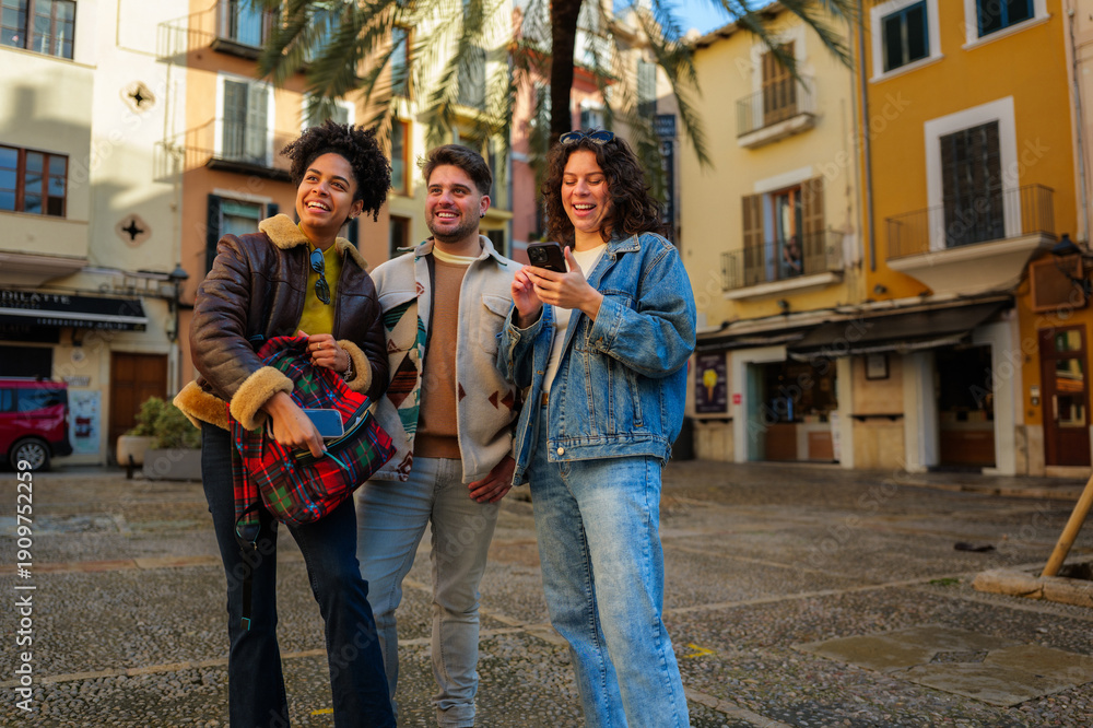 Obraz premium Diverse group of young adult friends enjoying urban travel, smiling and navigating a foreign city street together with a smartphone, finding directions while exploring