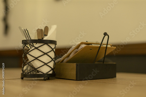 Metal napkin holder with tongs and bread tray on table, organized condiment and serving setup in cafe or bistro, neutral palette and focused shallow depth emphasizing texture and utility