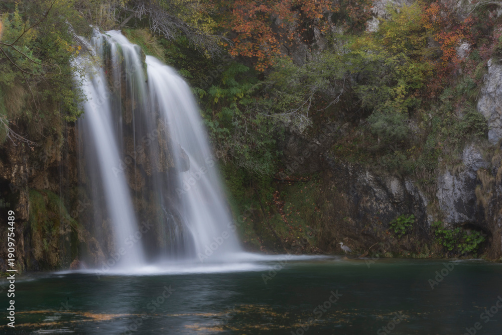 Obraz premium Professional long exposure of a waterfall cascading into a dark pond. Features autumn foliage, rocky textures, and a small heron. High-resolution nature photography for travel and editorial use.