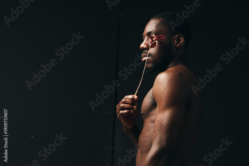 Принти на полотні Creative visual designer style portrait of dark skinned African American man holding dried flower near face with bare torso on black background in artistic concept