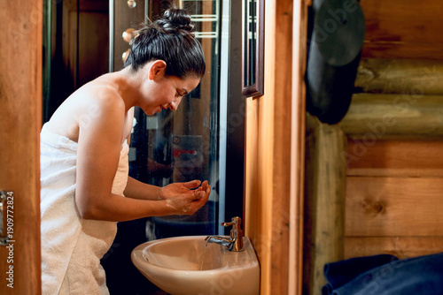 Obraz na plátne Woman in a towel cleansing her face at a bathroom sink in a rustic wooden cabin