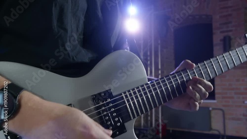 Closeup of caucasian young man in black t shirt playing white electric guitar on stage indoors with brick wall and stage lights, symbolizing energetic rock performance.