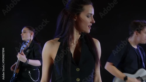 Full length view of caucasian young music band performing on stage indoors with bright round LED lights, female singer singing and musicians playing electric guitar and bass guitar.