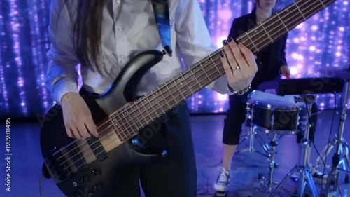 Closeup of caucasian young woman in white shirt and black bow tie playing black bass guitar on stage indoors with purple LED background, symbolizing live performance and music band energy.