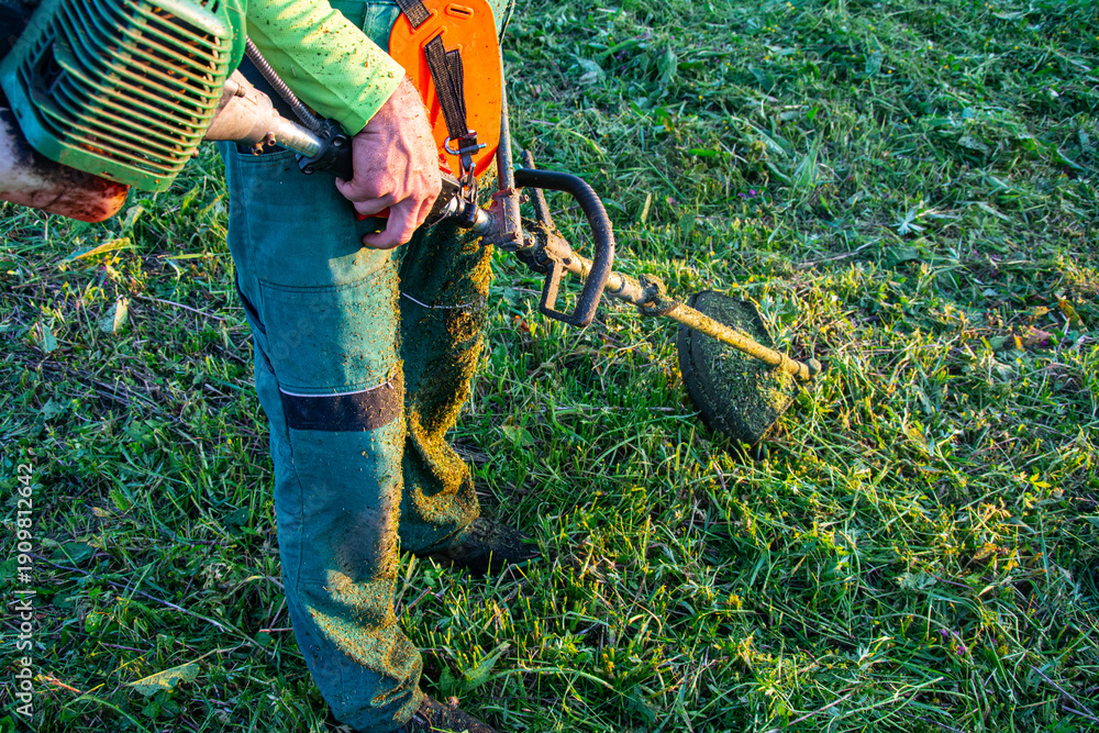 Obraz premium Worker Cutting Grass with Brush Cutter at Sunset