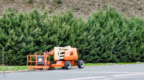 Tableau sur toile Orange articulated boom lift parked on a roadside against a green hedge