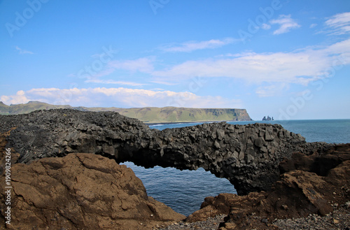 Basalt sea arch at Dyrhólaey on the south coast of Iceland overlooking the North Atlantic, with rugged volcanic cliffs and distant sea stacks under a blue sky.