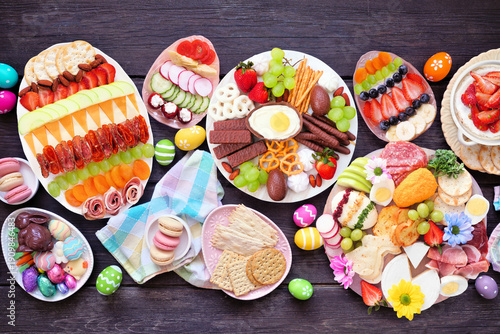 Spring or Easter grazing board theme table scene against a dark wood background. Top down view. Selection of sweets and cheese, meat and fruit appetizers.