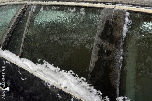 Car Window Covered in Thick Ice After Winter Storm. A close‑up view of a car door and window completely covered in a thick layer of ice and snow after a severe winter storm. Frozen glass, icy texture.