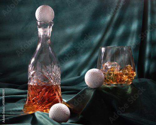 Glass of whiskey with ice cubes and golf ball on green satin background