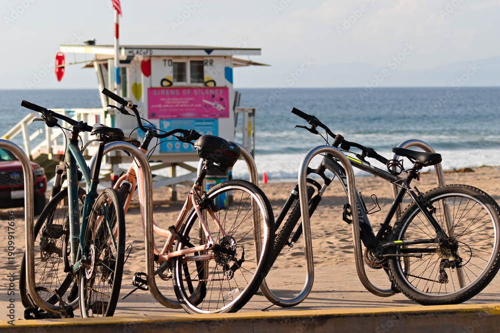 Fototapeta premium Bicycles secured to rack on sandy California beach with lifeguard tower and ocean waves in background.