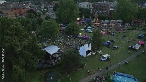 An aerial view shows a lively outdoor fairground event in a park, with carnival rides, food stalls, a stage, and a large crowd enjoying the festivities.