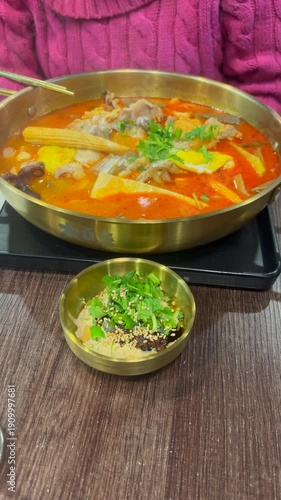 Vertical shot of a person from chest down, in winter clothing, prepared to eat an individual hot pot dish at a restaurant. Plastic tray underneath the metal shallow pot, with a side dish of sauce.