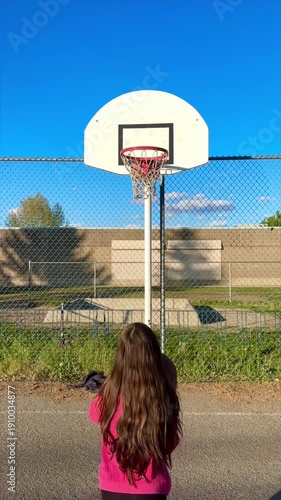 Vertical shot of a young girl with long curly brown hair, shooting a basketball outdoors and making the shot. 