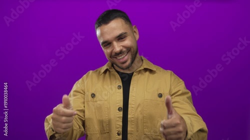 Man wearing tan jacket smiles and points finger to shirt while giving thumbs up in purple studio; confidence.