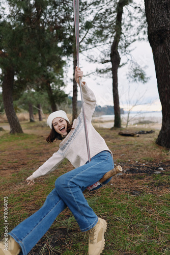 Girl on a swing in a park, outdoor joyful moment with trees and grass, smiling casually in a cozy sweater, jeans and boots, capturing playful motion and carefree childhood energy.