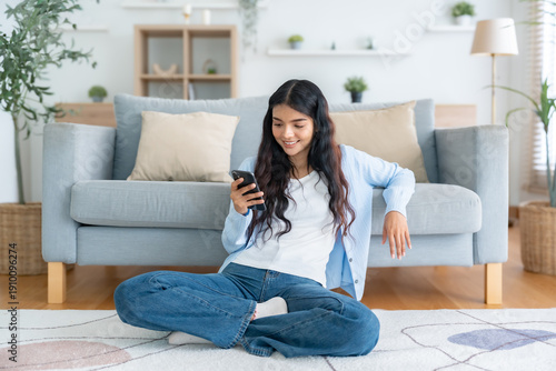 Smiling pretty young Asian woman sitting on the floor, using funny mobile apps in living room. Woman at Home, Doing Online Shopping, Messaging Friends, Posting on Social Media