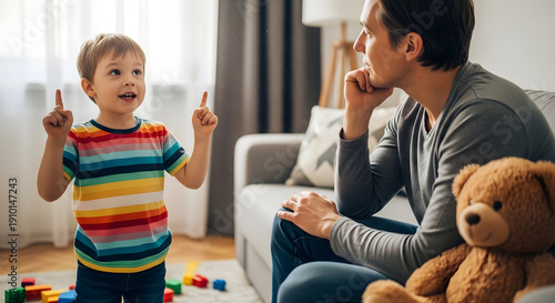 A man sitting on a sofa watching his toddler son talk and gesture with toys scattered on the floor