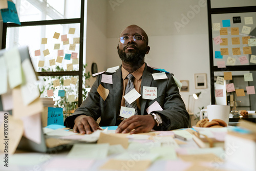 Schilderij op canvas Black man sitting at cluttered office desk covered with sticky notes, wearing gl