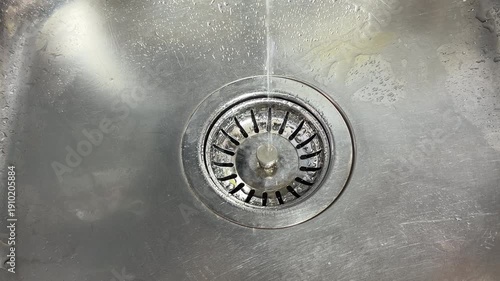 A thin stream of water falls into a metal sink basin. The liquid strikes the stainless steel drain, creating a delicate and rhythmic tinkling sound.
