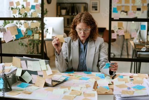 Quadro su tela Caucasian woman sitting at cluttered desk covered with sticky notes, holding not