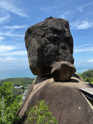 Massive granite rock formation perched on a hillside overlooking lush tropical forest and turquoise ocean coastline under a blue sky.