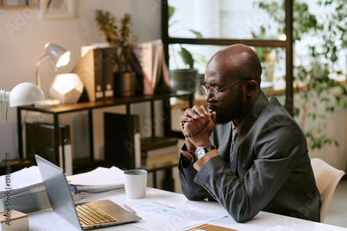 Black man wearing glasses sitting at desk praying in front of laptop screen, hands clasped in front of face, working in modern office environment with documents visible
