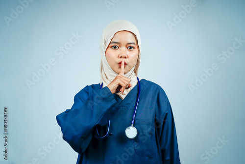 Serious Asian Muslim nurse or doctor in navy scrubs with a finger to her lips. Concept of patient privacy, medical confidentiality, quiet hospital zones, and data security.
