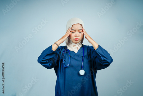 Muslim female nurse in hijab and blue scrubs holding her temples with both hands. Healthcare professional suffering from a headache, migraine, or high levels of stress.
