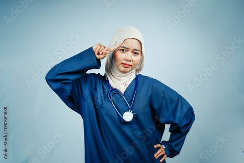 Serious Muslim female nurse in hijab and blue scrubs pointing a finger to her temple. Healthcare professional signaling intelligence, memory, or suggesting to 