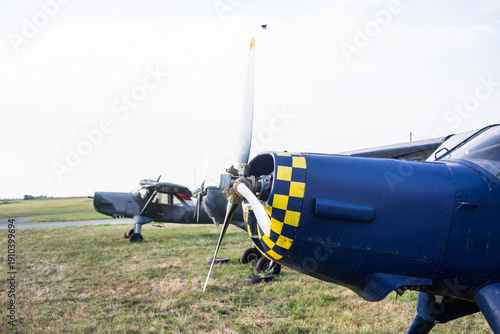 Close-up of the front of a blue piston plane with a yellow and black checkered pattern on the nose, parked on a grass runway.