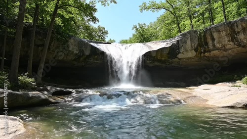 A majestic waterfall cascades from a rocky cliff, flowing into a clear, green pool surrounded by trees