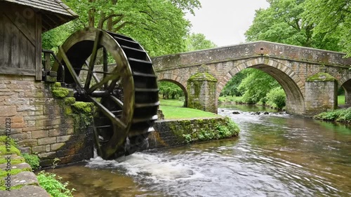 Wooden water wheel beside a stonework building and bridge over a flowing river