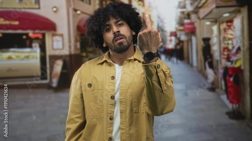 Man shows middle finger with both hands on a crowded street, curly hair and yellow jacket visible; urban defiance.